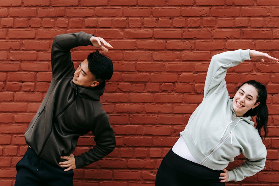 Positive young plus size female and fit Asian male trainer standing near brick wall and doing Overhead Side Reach Stretch exercise before workout on street